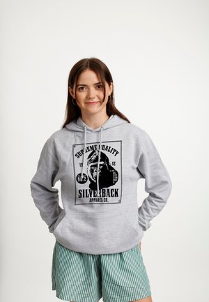 Jonge vrouw met lang donker haar die een grijze hoodie met een gorillagrafiek en gestreepte groene korte broek draagt, staand met haar handen achter haar rug.