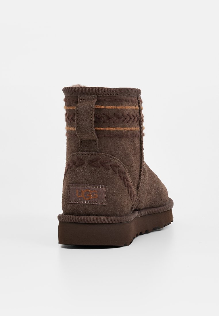 Bottines en suède marron avec un intérieur moelleux, des coutures décoratives et une étiquette à l'arrière. La semelle est plate et texturée pour un meilleur grip.