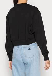 Žena v černé zkrácené mikině a světlých modrých džínech s vysokým pasem, na zadní kapse s malým černým trojúhelníkovým logem.