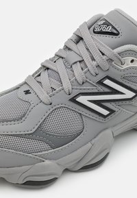 Chaussure de sport grise en matière synthétique et en maille, avec un bout rond, une semelle épaisse et une marque noire et blanche bien visible sur le côté.