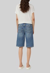 Shorts en denim avec une coupe décontractée, couleur bleu moyen et ourlet effiloché. Les poches arrière sont visibles, et un t-shirt blanc est porté par-dessus.