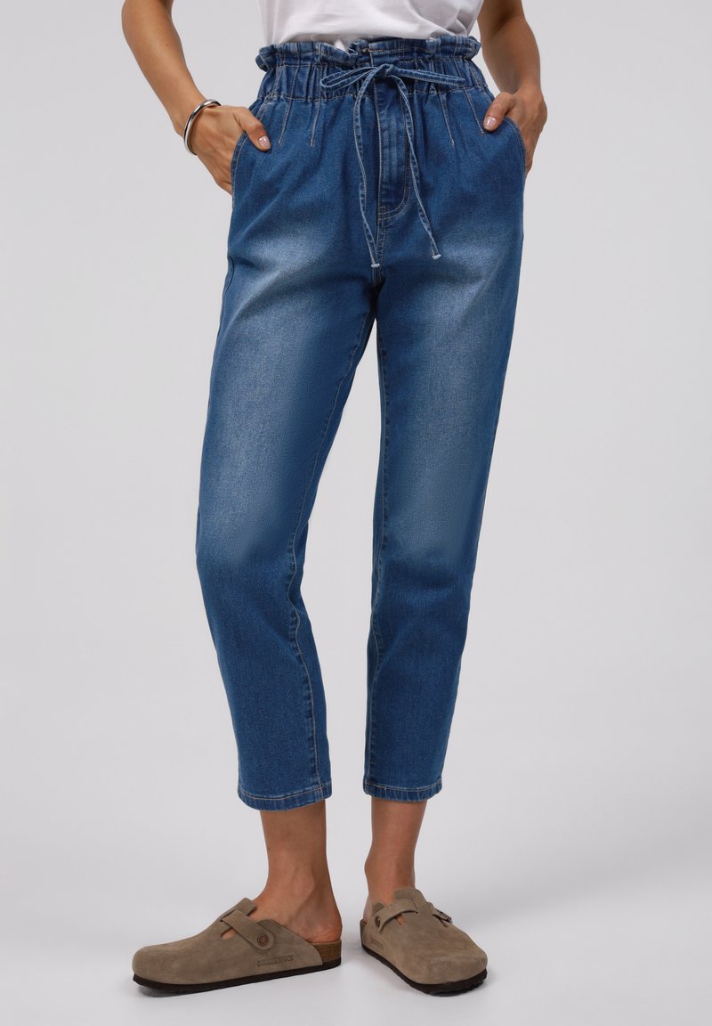 Femme portant un pantalon en denim bleu taille haute, fuselé, à taille paperbag avec cordon, associé à des sabots beiges à enfiler.