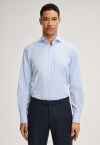 Camicia azzurro chiaro con maniche lunghe, parte anteriore con bottoni e colletto a punta; abbinata a pantaloni scuri. Tessuto liscio, vestibilità su misura.