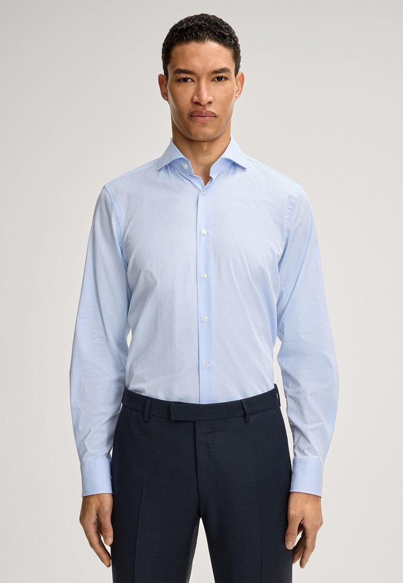Camicia azzurro chiaro con maniche lunghe, parte anteriore con bottoni e colletto a punta; abbinata a pantaloni scuri. Tessuto liscio, vestibilità su misura.