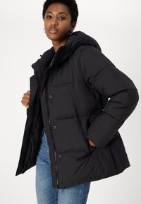 Black puffer jacket with a hood, featuring a zip front, snap buttons, and quilted texture. Model wearing it with blue jeans.