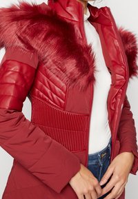 Chaqueta acolchada roja con cuello de piel sintética, secciones de tela acanalada y acolchada, diseño de cuello alto y cremallera frontal en una forma ajustada.
