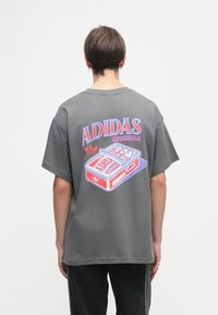 Pelēks kokvilnas T-krekls ar krāsainu grafiku uz muguras, uz kuras ir uzraksts "ADIDAS ORIGINALS" un bundžas dizains, brīvs piegriezums, īsas piedurknes.