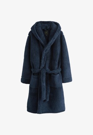 Homme aux cheveux courts remontant la capuche d’un manteau bleu foncé à texture duveteuse, regardant sur le côté sur un fond uni.