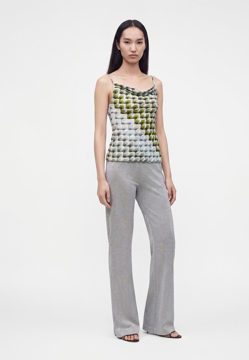 Femme portant un haut sans manches à motifs géométriques verts et bleus et un pantalon gris à jambes larges, debout devant un fond uni.