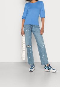 Modrá blůza s balónovými rukávy, roztrhané světle modré džíny s bílými skvrnami barvy, bílá ledvinka a vícebarevné tenisky s modrými akcenty.