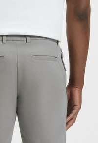 Pantalons gris avec une texture lisse, dotés de deux poches arrière et d'un zip sur le côté droit. Portés avec un haut blanc.
