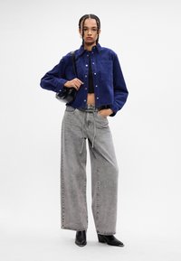Giacca in corduroy blu navy, corta con una tasca, abbinata a pantaloni jeans grigi larghi e stivaletti neri, mentre tiene una piccola borsa nera.
