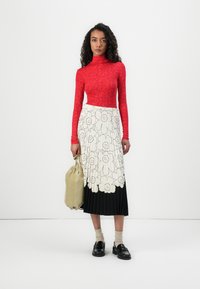 Rotes langärmliges Oberteil mit floralem Muster; weiße plissee Maxiröhre mit schwarzen Akzenten; kurze Socken und schwarze Loafer; hält eine grüne Tasche.