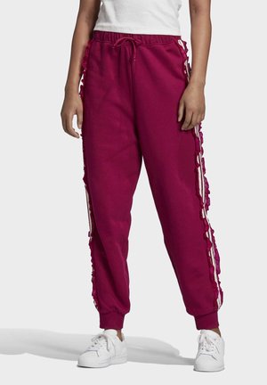 Personne portant un pantalon de jogging bordeaux avec un ourlet à volants et des rayures blanches, assorti à des baskets blanches et un haut blanc.
