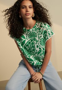 Green and white patterned T-shirt with floral designs and scalloped sleeves, paired with light blue denim jeans.