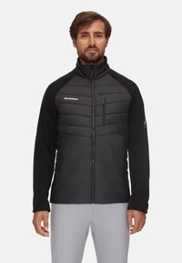 Hombre con una chaqueta de cremallera negra de Mammut, con parte frontal acolchada y mangas de forro polar, de pie contra un fondo claro y liso.