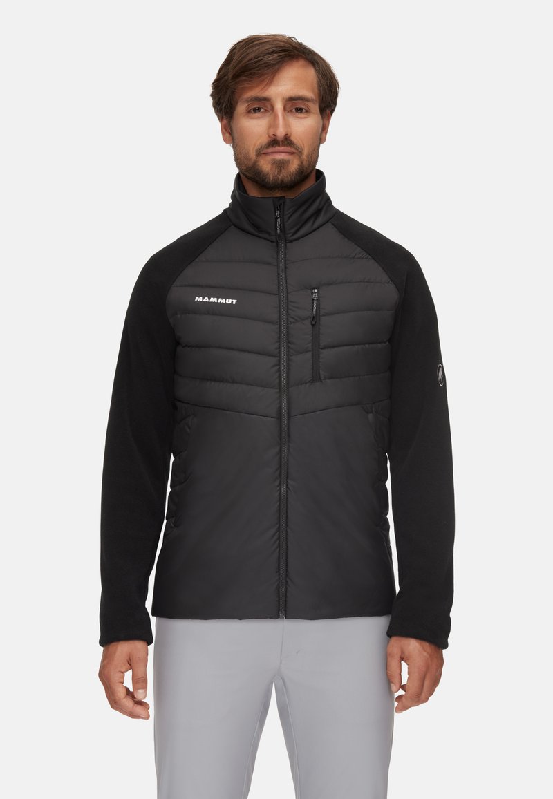 Hombre con una chaqueta de cremallera negra de Mammut, con parte frontal acolchada y mangas de forro polar, de pie contra un fondo claro y liso.