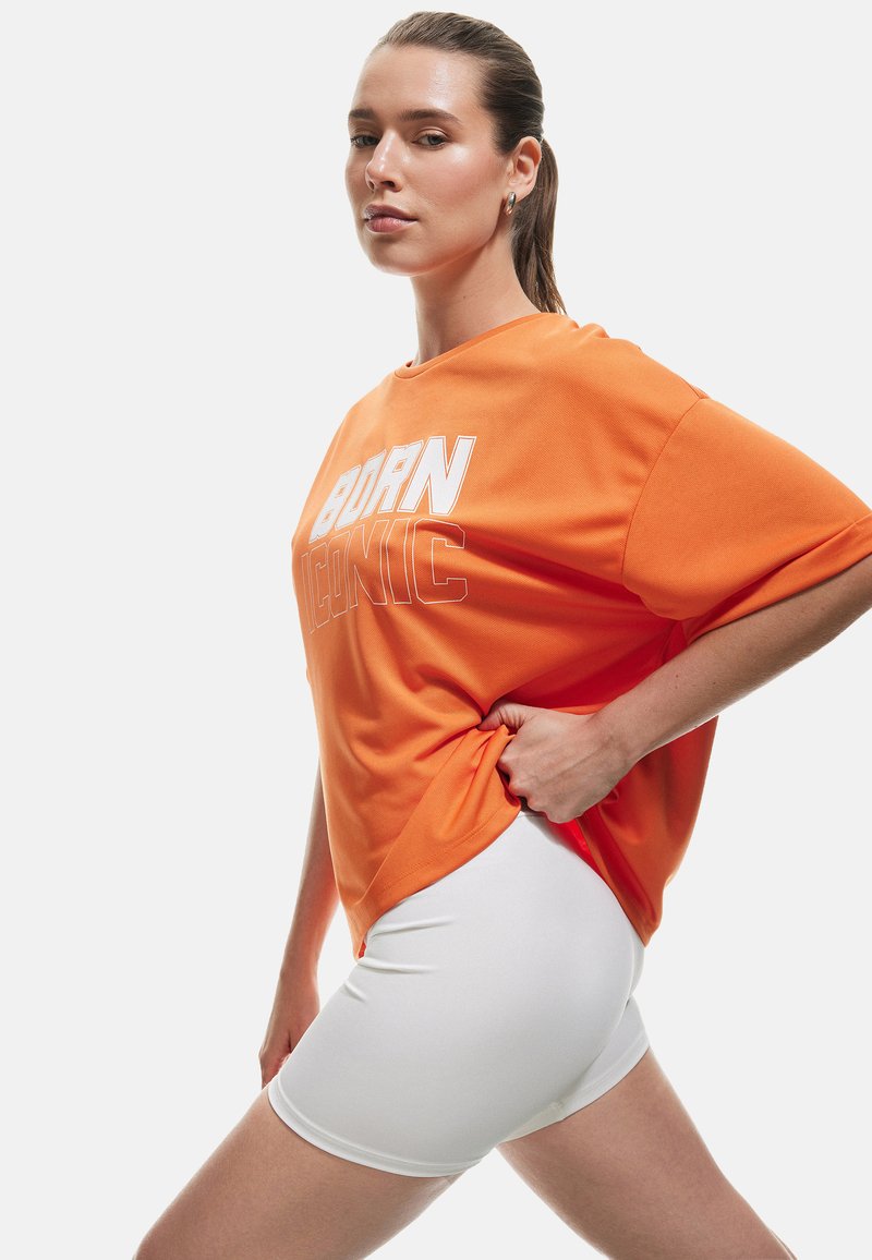 Camiseta naranja de gran tamaño con "BORN ICONIC" impreso en blanco, combinada con pantalones cortos ajustados blancos, contra un fondo blanco liso.