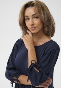 Navyblaues, strukturiertes Kleid mit Puffärmeln und gebundenen Akzenten an den Manschetten. Model posiert mit der Hand am Kinn, neutraler Hintergrund.