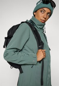 Grønn softshell-jakke med høy krage, glidelås og justerbare funksjoner, brukt over en svart ryggsekk, kombinert med en lue og solbriller.