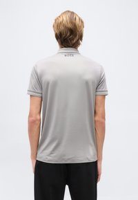 Ξανθός/ή φοράει κοντομάνικη πόλο σε ανοιχτό γκρι με την επιγραφή "BOSS" στον πίσω γιακά, συνδυασμέν η με μαύρο παντελόνι, στέκεται μπροστά σε απλό φόντο.