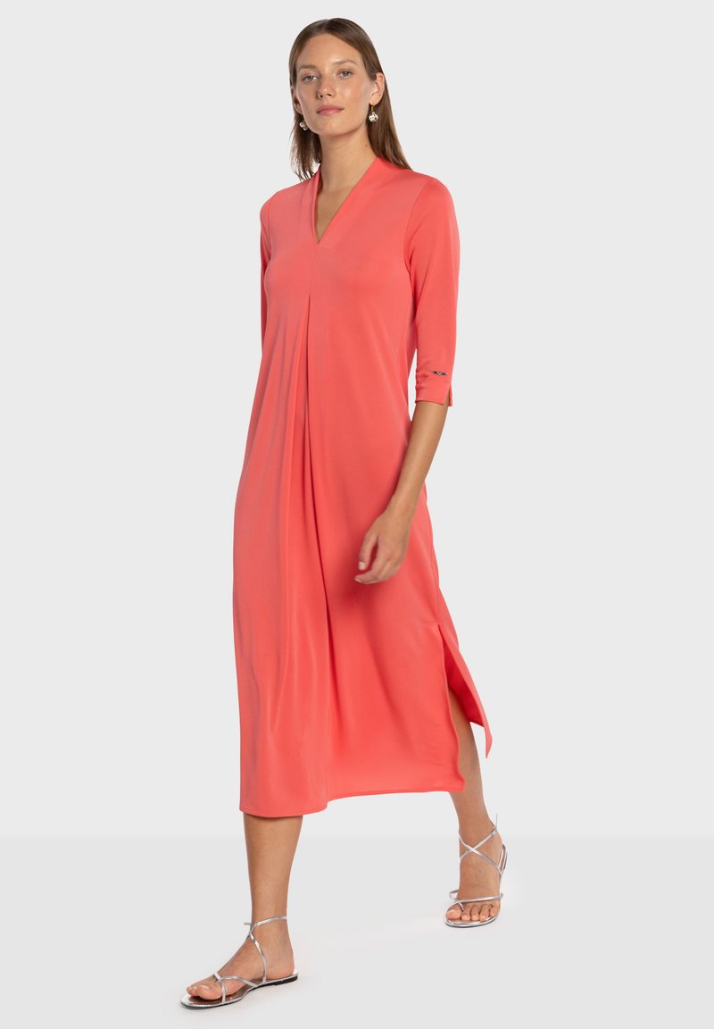 Koralfarvet, midilengde kjole lavet af et blødt, glat stof. Har en v-udskæring, trekvartærmer og sidelommer for nem bevægelse.