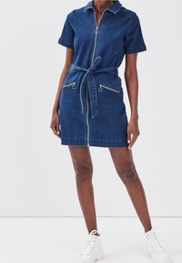 Robe en jean à manches courtes, fermeture éclair frontale, ceinture à la taille et deux poches zippées ; couleur bleu foncé avec détails de couture blanche.