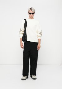 Witte lange mouw shirt met grafische details, zwarte cargobroek en grijze sneakers. Het model draagt een zwarte tas en heeft een zonnebril op.