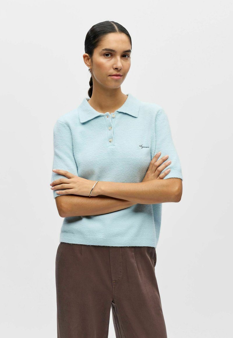 Polo à manches courtes bleu clair en tissu doux et texturé. Présente trois boutons et un petit logo sur le côté gauche.