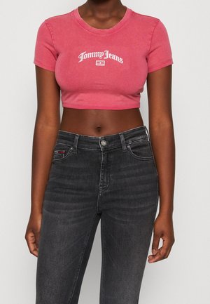 Kvinde iført en tætsiddende, rød Tommy Jeans crop top og højtaljede mørkegrå jeans, stående mod en ensfarvet lys baggrund.