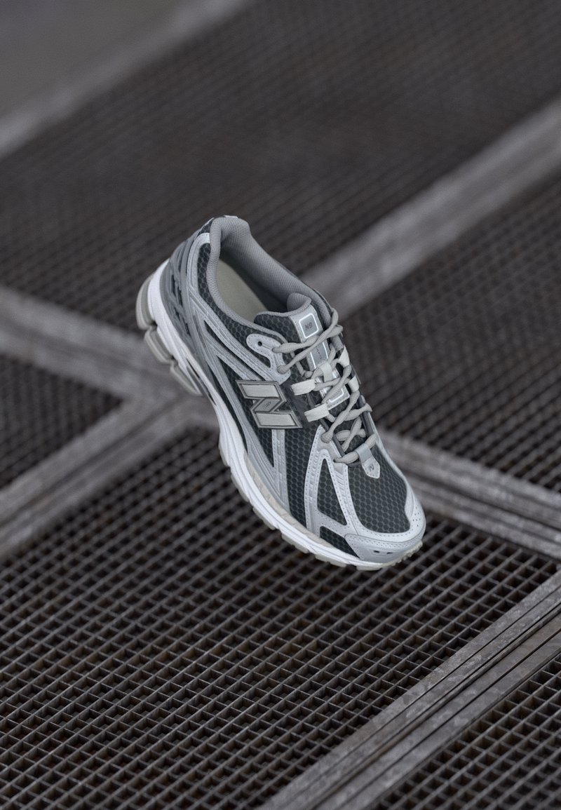 Gray and white New Balance running shoe with laces on metal grid surface with cross pattern.