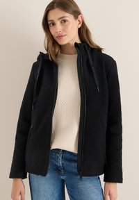 Veste en polaire noire avec un devant zippé, une capuche doublée et des poches zippées ; le tissu a une texture douce, complété par un pull en tricot côtelé.
