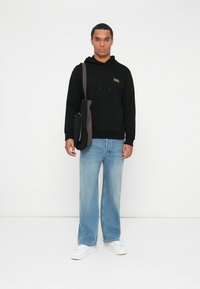 Sudadera negra con logo dorado, vaqueros de pierna ancha en azul claro y zapatillas blancas. Bolso negro de hombro con detalle de cremallera.