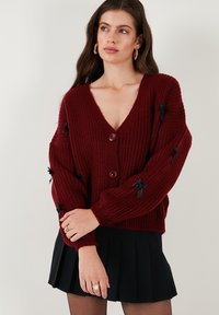 Burgundy ullblandad cardigan med djup V-hals, knappstängning och svarta rosettaccenter på ärmarna, med ribbad textur.