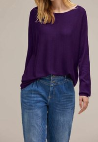 Kvinna i en löst sittande lila långärmad topp instoppad i högmidjade blå jeans med synliga knappar och veck.