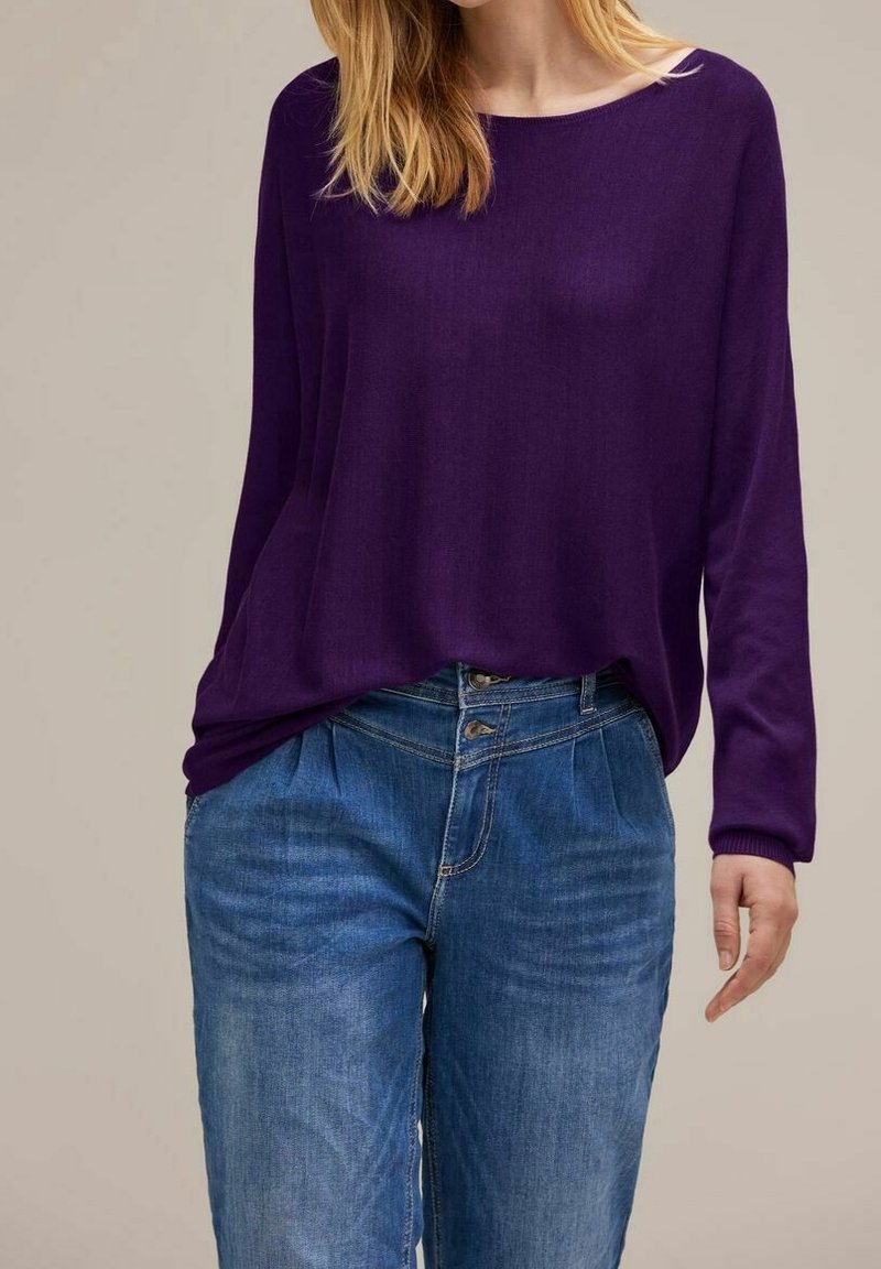 Kvinna i en löst sittande lila långärmad topp instoppad i högmidjade blå jeans med synliga knappar och veck.