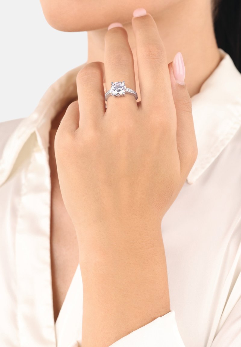 Kvinnas hand nära ansiktet som visar en silverförlovningsring med stor rund diamant och mindre sidostenar, bärande en vit skjorta.
