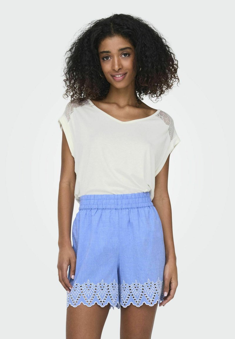 Luźna kremowa bluzka z dekoltem w serek i koronkowymi detalami na ramionach, zestawiona z jasnoniebieskimi szortami z białym wykończeniem w formie oczek na dolnej krawędzi.
