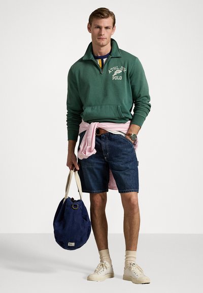 Felpa verde scuro con colletto zip e testo "ATHL. DIV. 67 POLO", pantaloni corti di jeans, camicia rosa legata in vita e borsa di tela blu. Sneakers bianche.