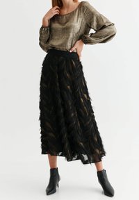 Metalická bronzová blůza s dlouhým rukávem spárovaná s černou midi sukní s volány, která má zlaté detaily a texturované třásně. Černé kotníkové boty.