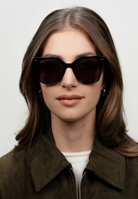 Černé oversize sluneční brýle s lesklým povrchem, úhlovým designem a kovovými akcenty na rámu. Model má na sobě tmavou bundu.