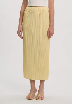 Vrouw draagt een hooggetailleerde, geplooide gele midi-rok met een dunne strikceintuur, gecombineerd met gele sandalen met hak en een wit cropped topje.