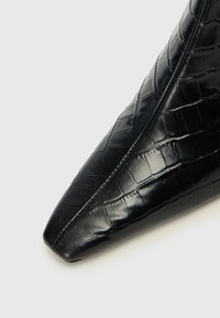 Chaussure noire en cuir verni à bout pointu avec une texture crocodile et un design épuré, présentant une finition lisse et des détails de couture minimalistes.