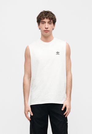 Młody mężczyzna o kręconych włosach, ubrany w białą bezrękawnikową koszulkę Adidas i czarne spodnie cargo, stojący na jasnym, jednolitym tle.