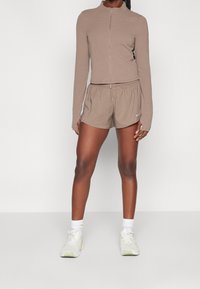 Woman wearing taupe zip-up long-sleeve top and matching athletic shorts, white socks, and white running shoes, standing on a plain background.