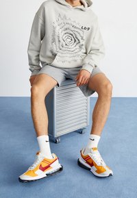 Sweat à capuche gris avec motif graphique et texte, short gris, et chaussettes blanches Nike. Les chaussures présentent un mélange de couleurs orange, rouge et blanc.
