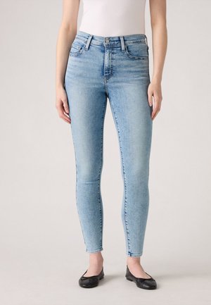 Jean skinny bleu clair en denim, avec taille haute, cinq poches et décoloration subtile. Porté avec des ballerines noires.