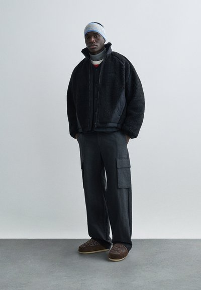 Svart shearlingjacka med hög krage, lager-på-lager över en grå tröja, kombinerad med mörka cargobyxor och bruna broderade slip-on-skor.