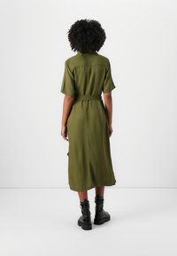 Abito midi verde oliva con maniche corte, vita cintrata, texture liscia e dettaglio con spacco sul retro, abbinato a stivali neri alla caviglia.