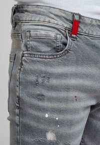 Gros plan sur un jean en denim gris avec des zones usées, des éclaboussures de peinture blanche et une petite étiquette rouge sur la boucle de la ceinture.